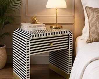 May include: A bedside table featuring a black and white striped pattern with a single drawer, a small gold knob, and gold-toned base accents. The table is topped with a white dome-shaped lamp and two books titled Live Beautifully and This Is Home.
