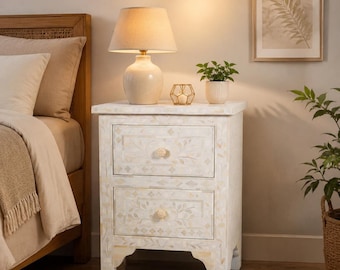 May include: A white bedside table featuring two drawers with intricate mother-of-pearl inlay patterns, floral motifs, and round knobs. The rectangular piece stands on four short legs and displays a smooth, light-colored finish throughout.