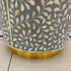 May include: A cylindrical side table featuring a dark gray surface inlaid with a repeating white and cream leaf pattern made of mother-of-pearl. The base of the table is constructed from a smooth, polished gold-toned metal band.