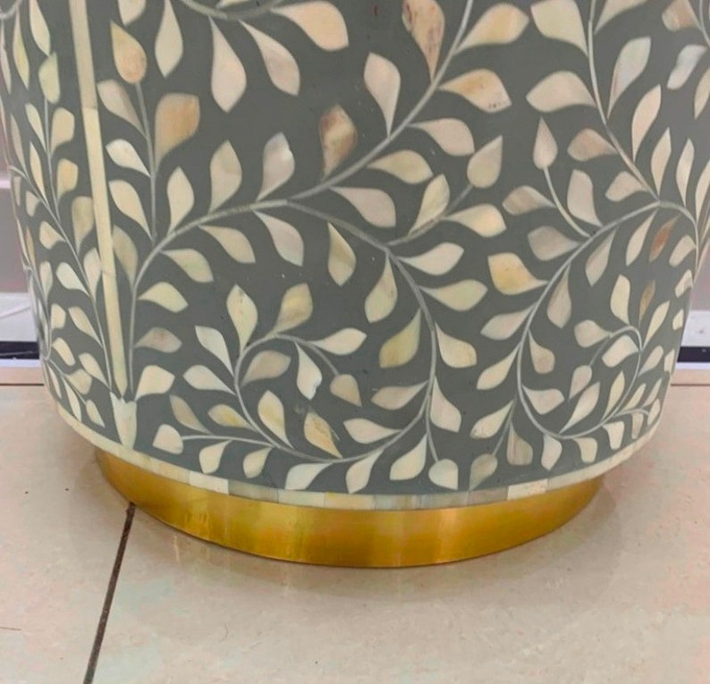 May include: A cylindrical side table featuring a dark gray surface inlaid with a repeating white and cream leaf pattern made of mother-of-pearl. The base of the table is constructed from a smooth, polished gold-toned metal band.