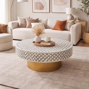 May include: A circular coffee table with a patterned surface and a gold base is the focal point of a living room. The table is surrounded by a cream sofa, cushions, and a rug. Decorative items are on the table.