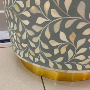 May include: A cylindrical side table featuring a grey base with an intricate inlaid pattern of cream-colored leaf shapes. The bottom of the table is finished with a smooth, reflective gold-toned metal band that encircles the base of the furniture.