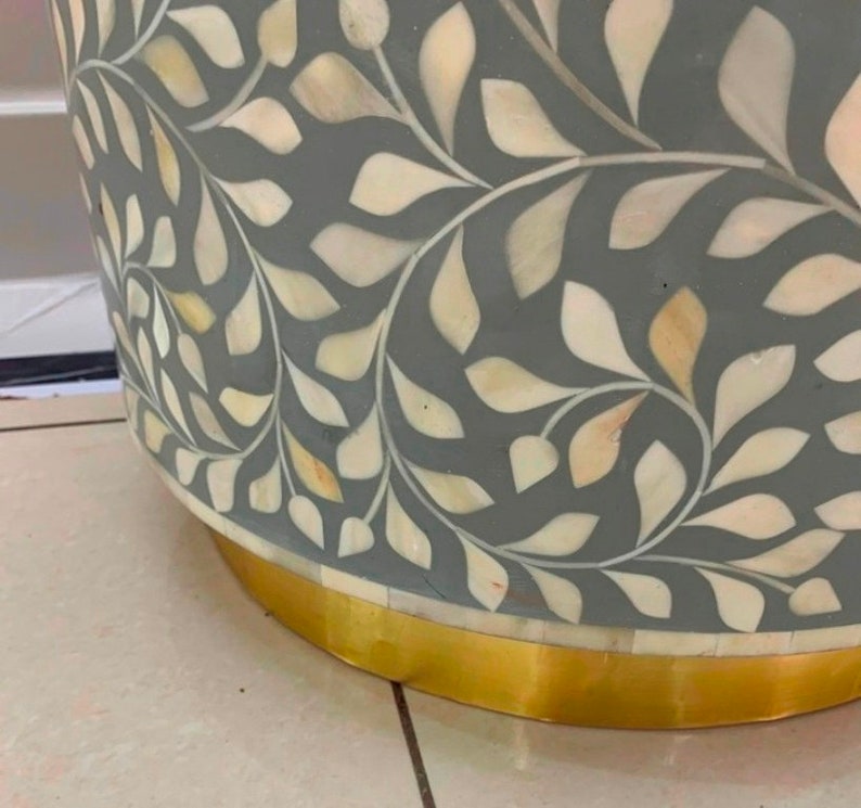 May include: A cylindrical side table featuring a grey base with an intricate inlaid pattern of cream-colored leaf shapes. The bottom of the table is finished with a smooth, reflective gold-toned metal band that encircles the base of the furniture.
