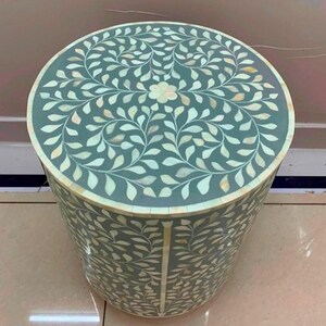 May include: A cylindrical side table featuring a sage green base with an intricate inlaid pattern of white, leaf-shaped pieces. The top surface displays a symmetrical floral and vine motif, while the sides are covered in a repeating leaf design.