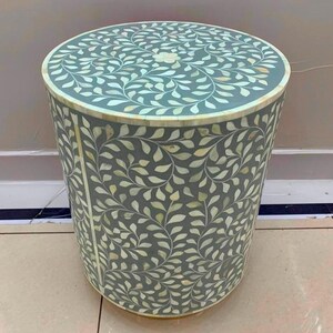 May include: A cylindrical side table featuring a grey base with an intricate, repeating white leaf pattern inlaid across the top and sides. The design includes a central floral motif on the circular top surface and a smooth, uniform finish.