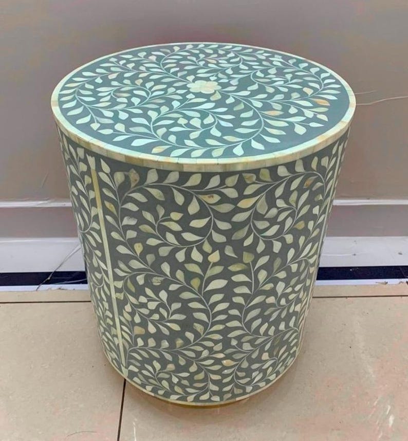 May include: A cylindrical side table featuring a grey base with an intricate, repeating white leaf pattern inlaid across the top and sides. The design includes a central floral motif on the circular top surface and a smooth, uniform finish.