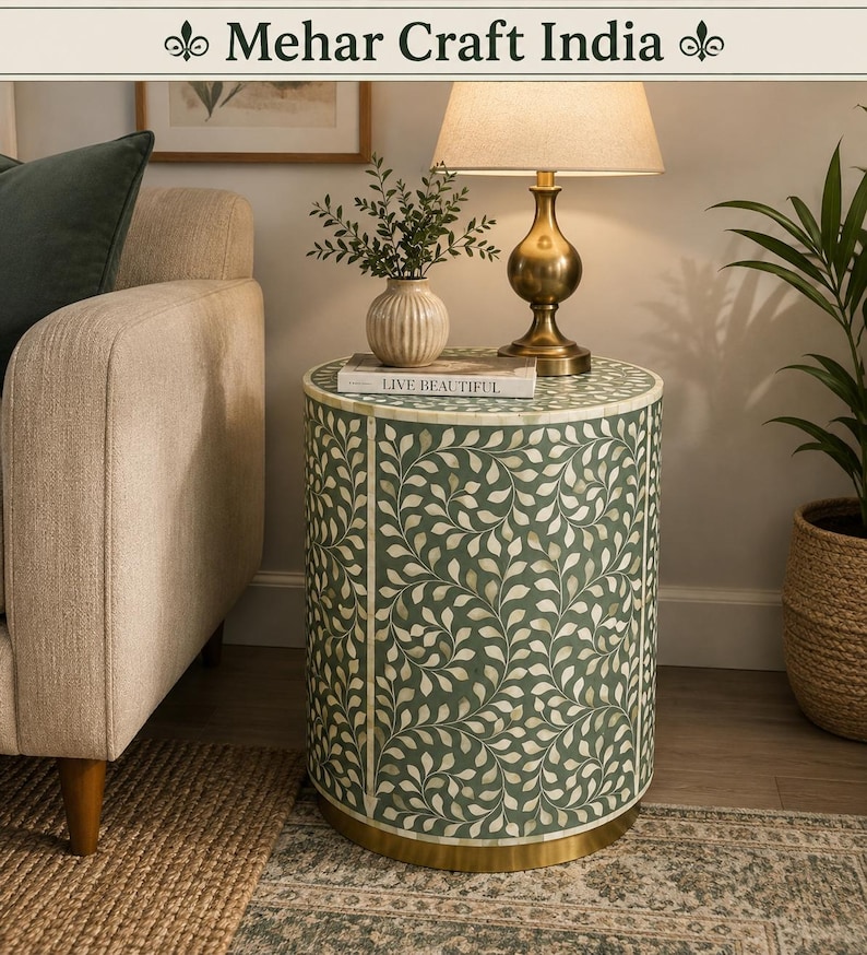 May include: A cylindrical side table featuring a sage green surface with an intricate white vine and leaf inlay pattern. The table has a solid gold-toned metal base and a smooth, flat top surface. The text Mehar Craft India is visible at the top.