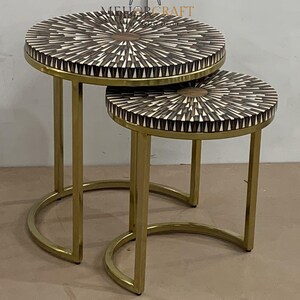 Puede incluir: Un conjunto de dos mesas nido de metal dorado con tapas redondas. Las tapas son de madera con un patrón geométrico en tonos marrones y blancos.