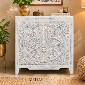 Op de afbeelding: Een rechthoekige houten kast met twee deuren voorzien van ingewikkelde, met de hand gesneden bloem- en cirkelpatronen. De kast heeft een verweerde witte afwerking met zichtbare blauwe ondertonen, een plat bovenblad en vier korte, stevige poten.