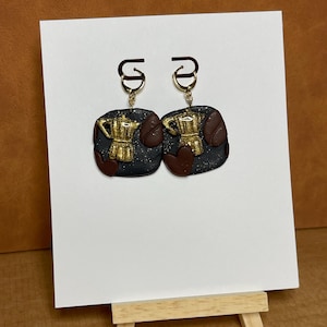 May include: A pair of handmade earrings featuring a coffee theme. Each earring has a black base with gold-colored coffee maker and brown coffee bean accents. The earrings hang from gold-colored ear wires.
