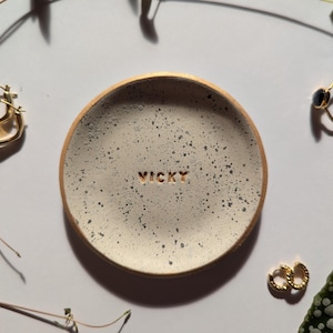 Peut inclure: Un plat à anneaux en céramique blanche avec une finition mouchetée et le nom "VICKY" inscrit en lettres dorées. Le plat est posé sur une surface blanche avec des boucles d'oreilles dorées et une bague.