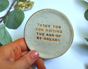 May include: A small, white ceramic dish with a speckled finish. The dish has a gold inscription that reads "Thank you for raising the man of my dreams".