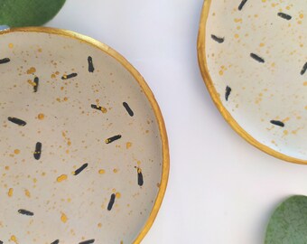 Puede incluir: Dos platos decorativos pequeños, redondos, con base blanca, borde dorado y diseño abstracto en negro y dorado.