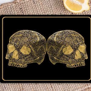 Puede incluir: Dos diseños de calaveras de color dorado que se miran en un fondo negro. Las calaveras están detalladas con intrincados patrones.
