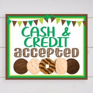 Peut inclure: Un panneau brun et vert avec le texte "CASH & CREDIT accepted" en lettres vertes. Il y a cinq biscuits Girl Scout en rangée au bas du panneau.