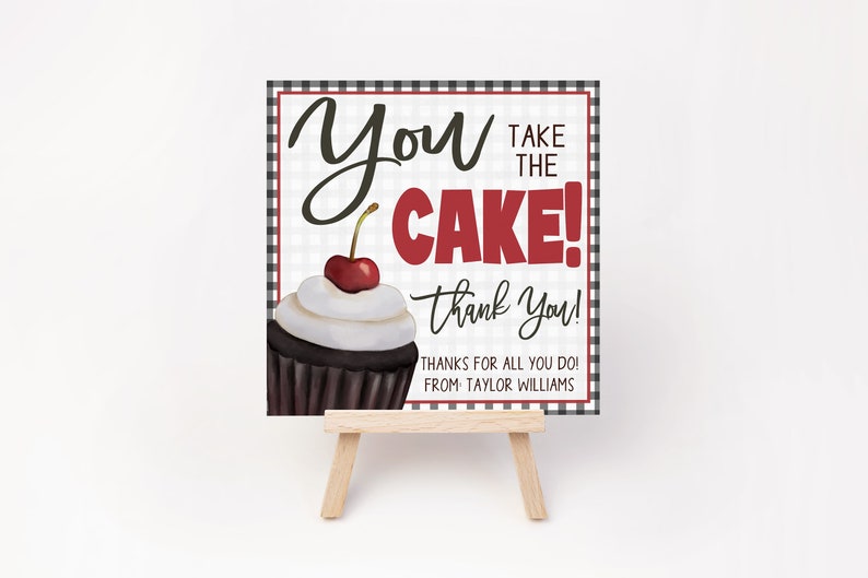 May include: A black and white checkered sign with the text "You Take the Cake! Thank You!" in red and black. A chocolate cupcake with white frosting and a red cherry is pictured on the sign. The sign is on a small wooden easel.