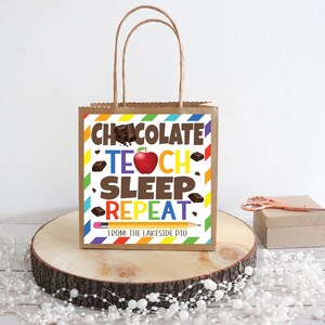 Puede incluir: Una bolsa de regalo de papel marrón con un diseño a rayas de colores y una etiqueta blanca que dice "Chocolate Teach Sleep Repeat" con una manzana roja y trozos de chocolate. La etiqueta también dice "De: The Lakeside PTO".