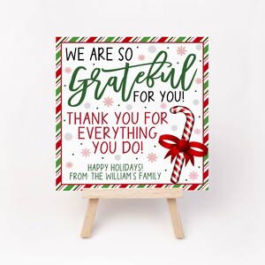 May include: A white sign with red and green stripes around the border. The sign says "We are so Grateful for you! Thank you for everything you do! Happy Holidays! From the William's Family". A candy cane with a red bow is in the bottom right corner of the sign.