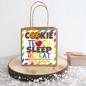 Puede incluir: Una bolsa de regalo de papel marrón con un diseño de rayas de colores y una etiqueta blanca que dice "Cookie Teach Sleep Repeat" con un gráfico de una galleta y una manzana. La etiqueta también dice "De: The Lakeside PTO".