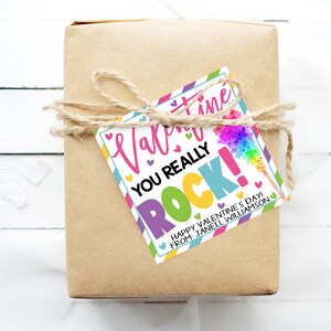 May include: A colorful Valentine's Day gift tag with the text "Valentine's Day You Really Rock!" and a rainbow-colored cotton candy stick. The tag is tied with twine to a brown gift box. The text "Happy Valentine's Day from Janell Williamson" is also on the tag.