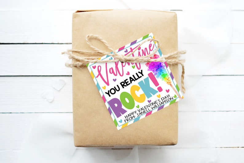 May include: A colorful Valentine's Day gift tag with the text "Valentine's Day You Really Rock!" and a rainbow-colored cotton candy stick. The tag is tied with twine to a brown gift box. The text "Happy Valentine's Day from Janell Williamson" is also on the tag.