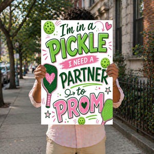 Può includere: Un cartello bianco con la scritta "I'm in a PICKLE I need a PARTNER to PROM" in verde e rosa, con racchette e palline da pickleball. Il cartello è tenuto da una persona che indossa una camicia rosa e pantaloncini kaki.