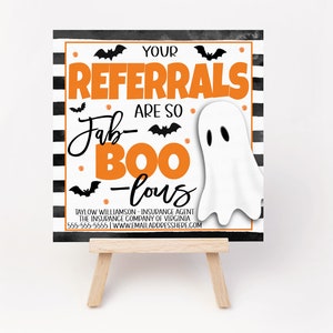 May include: A black and white striped sign with orange text that reads "Your Referrals Are So Fab-Boo-Lous". A white ghost with black eyes is on the sign. The sign is on a small wooden easel.
