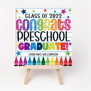 K&ouml;nnte beinhalten: Ein farbenfrohes Schild mit dem Text "Class of 2022 Congrats Preschool Graduate!" auf wei&szlig;em Hintergrund mit blauen Linien und Sternen. Das Schild steht auf einem kleinen Holzstaffelei. Der Text ist in einem Regenbogen von Farben. Am unteren Rand des Schildes befinden sich Buntstifte.