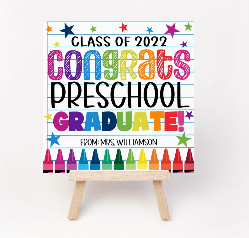 K&ouml;nnte beinhalten: Ein farbenfrohes Schild mit dem Text "Class of 2022 Congrats Preschool Graduate!" auf wei&szlig;em Hintergrund mit blauen Linien und Sternen. Das Schild steht auf einem kleinen Holzstaffelei. Der Text ist in einem Regenbogen von Farben. Am unteren Rand des Schildes befinden sich Buntstifte.