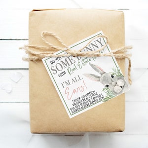 May include: A brown paper gift box tied with twine and a gift tag that says "Do you know some bunny with Real Estate needs? I'm all ears!" with a cartoon bunny illustration. The tag also includes contact information for a real estate agent.