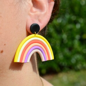 Puede incluir: Pendientes en forma de arcoíris con un disco negro en la parte superior. El arcoíris está hecho de arcilla polimérica amarilla, naranja, rosa, morado y blanco.