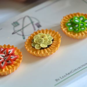 May include: Three miniature fruit tarts with different fillings. The first tart is filled with sliced strawberries, the second with sliced bananas, and the third with sliced kiwi. The tarts are on a white surface with the text "By LeaArtCollection" in the bottom right corner.