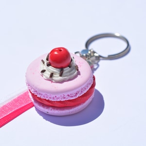 Könnte beinhalten: Ein rosa und roter Macaron-Schlüsselanhänger mit weissem Frosting und einer roten Kirsche obenauf. Der Schlüsselanhänger hat einen silbernen Ring.
