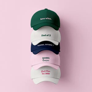 Peut inclure: Une pile de cinq casquettes de baseball de différentes couleurs : vert, blanc, bleu marine, rose et blanc. Chaque casquette porte une inscription blanche : "love wins.", "Dad of 3", "TENNIS, ANYONE?", "MAMMA love" et "Best Mom Est. 2022". Fond rose.