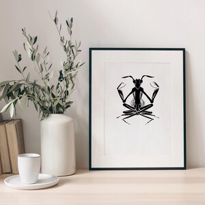 May include: A black and white framed art print featuring a stylized illustration of an insect in a seated yoga pose. The artwork is displayed on a wooden surface, next to a white vase with olive branches, books, and a cup.