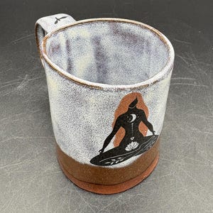 May include: Ceramic mug with a brown base and handle, featuring a white and purple speckled body. The mug has a black illustration of a person in a yoga pose with a crescent moon and floral design. The mug is suitable for hot or cold beverages.