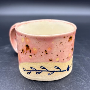 May include: Pink ceramic mug with a speckled glaze and a cream-colored interior. The mug features a blue leaf design and a small blue diamond shape near the base. The handle is also pink.