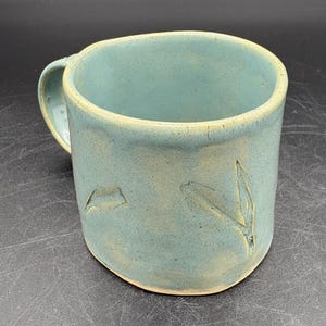 May include: A handmade ceramic mug with a light blue glaze. The mug features a rounded handle and leaf-like designs carved into the side. The interior and rim have a lighter, natural color.