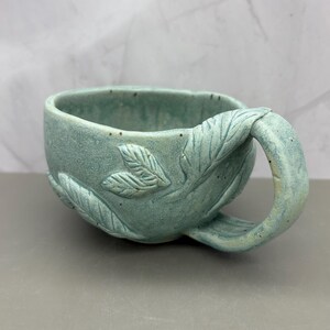 May include: A handmade, turquoise ceramic mug with a leaf design. The mug features a sculpted handle and leaf details on the body. The mug is approximately 4 inches wide and 3 inches tall.