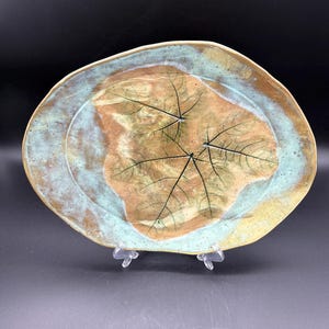 May include: A handmade ceramic serving platter with a leaf imprint design. The platter features a brown center with black leaf veins, surrounded by a turquoise and yellow rim. The organic shape and unique glaze give it a rustic, artistic feel.