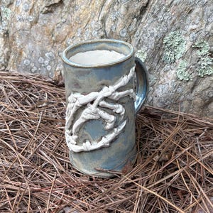 May include: A blue ceramic mug with a white bone-like design wrapped around the exterior. The mug has a handle and a textured surface. The background features a rock and dried pine needles.