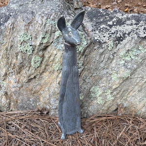 May include: A tall, gray metal sculpture of a rabbit with long ears. The rabbit statue is standing upright with its front paws together. The sculpture is set against a large rock with green and gray lichen.