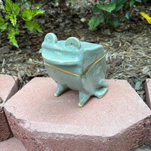 May include: A ceramic frog-shaped trinket box in a muted teal color with gold accents. The frog has a square body, small legs, and large eyes. The box is sitting on a red brick surface, with green foliage in the background.