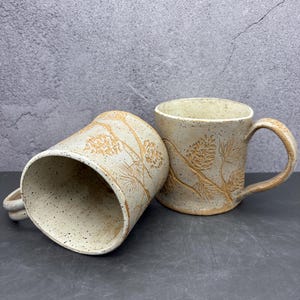 May include: Two speckled ceramic mugs with a nature-inspired design. The mugs are off-white with a brown pinecone and branch pattern. One mug is upright, and the other is tilted on its side, showcasing the interior.