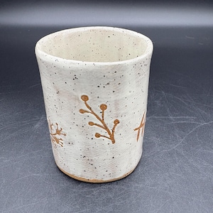 May include: A speckled ceramic tumbler with a wide opening. The off-white cup features brown floral designs and dark speckles. The bottom of the cup is a darker brown. The cup is approximately 4 inches tall.