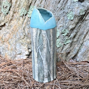May include: A cylindrical ceramic vase featuring a hand-painted forest scene with white birch trees against a muted gray background. The top rim of the vase is finished with a smooth, glossy light blue glaze that dips into a V-shape.