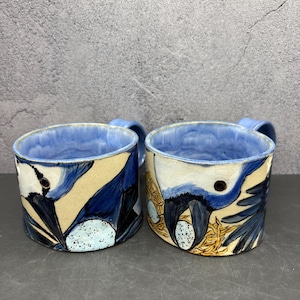 May include: Two ceramic mugs with blue interiors and handles. The exterior features a design of a bird with blue and black feathers, a tan background, and a nest with eggs. The mugs are approximately 4 inches tall.