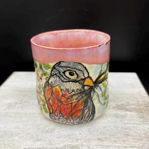 May include: A ceramic mug with a hand-painted bird design. The mug features a pink rim and a detailed illustration of a bird with orange, gray, and white feathers. The bird is set against a backdrop of green foliage and a light blue background.