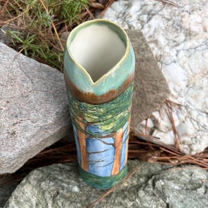 May include: A cylindrical ceramic vase featuring a hand-painted forest scene with tall brown tree trunks, green foliage, and a light blue sky. The rim has a distinct V-shaped notch and is glazed in a smooth, glossy sage green and brown finish.