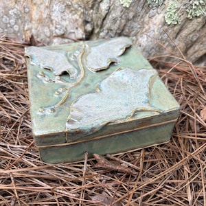 May include: A square ceramic box with a lid, featuring a textured, light green surface with raised, leaf-like designs in a lighter hue. The box has a gold-colored trim and is set on a bed of pine needles.